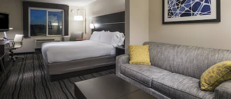 Habitación de hotel con cama, sofá gris con almohada amarilla, una mesita de café, escritorio con silla, televisión de pantalla plana y un cuadro abstracto en la pared.