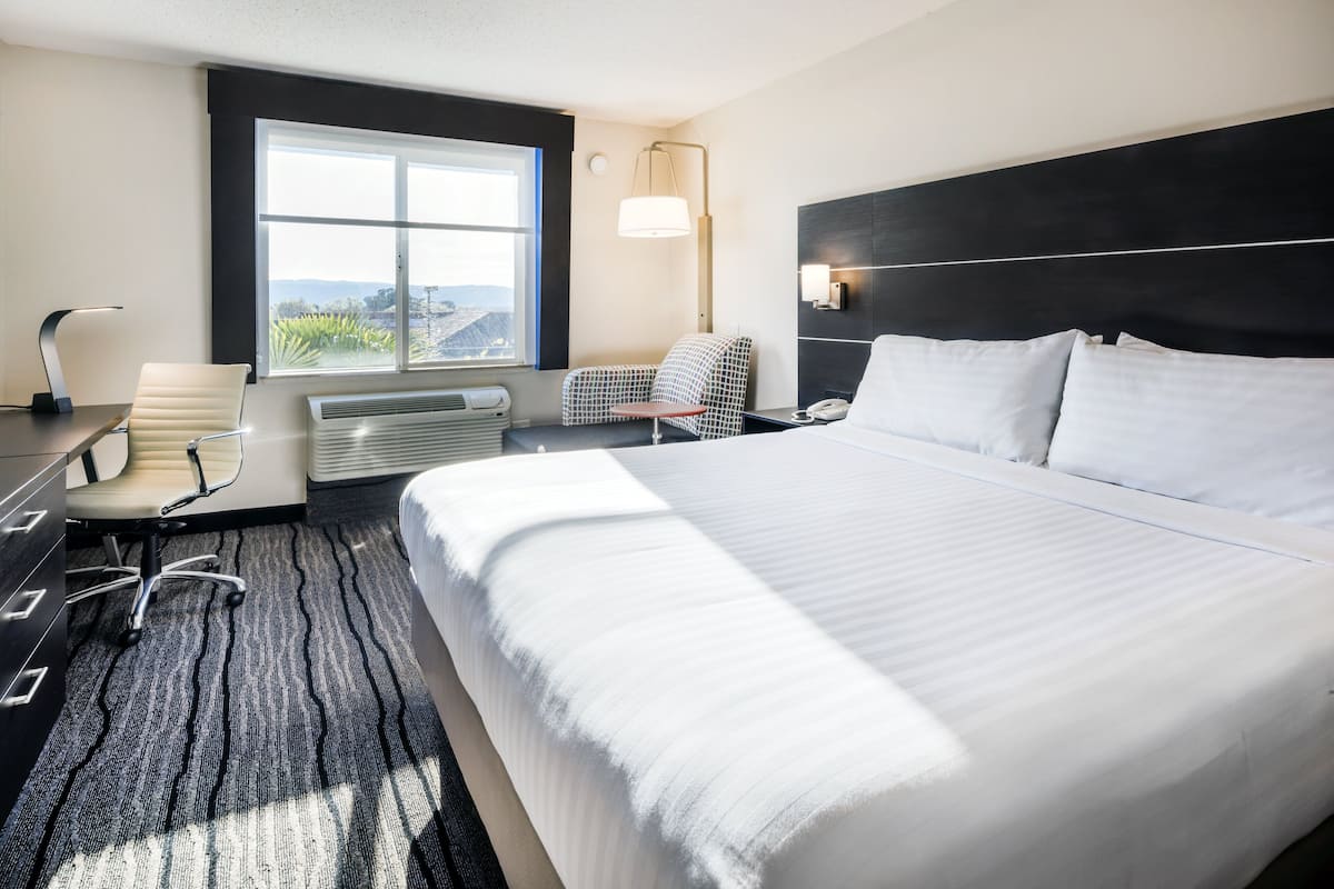 A modern hotel room with a large bed, a desk with a chair, a small table, and a window with a view.