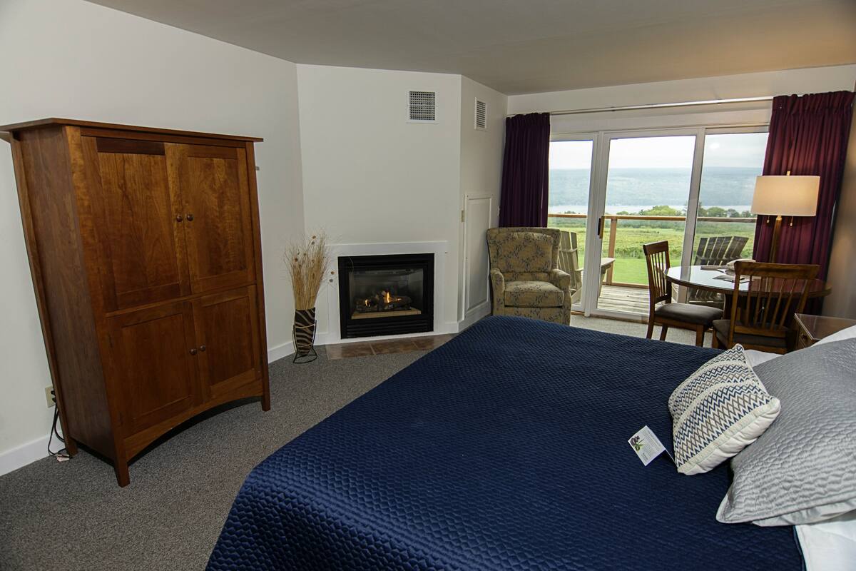 A hotel room with a large bed, a wooden wardrobe, a fireplace, a balcony with chairs, and a view of the outdoors.
