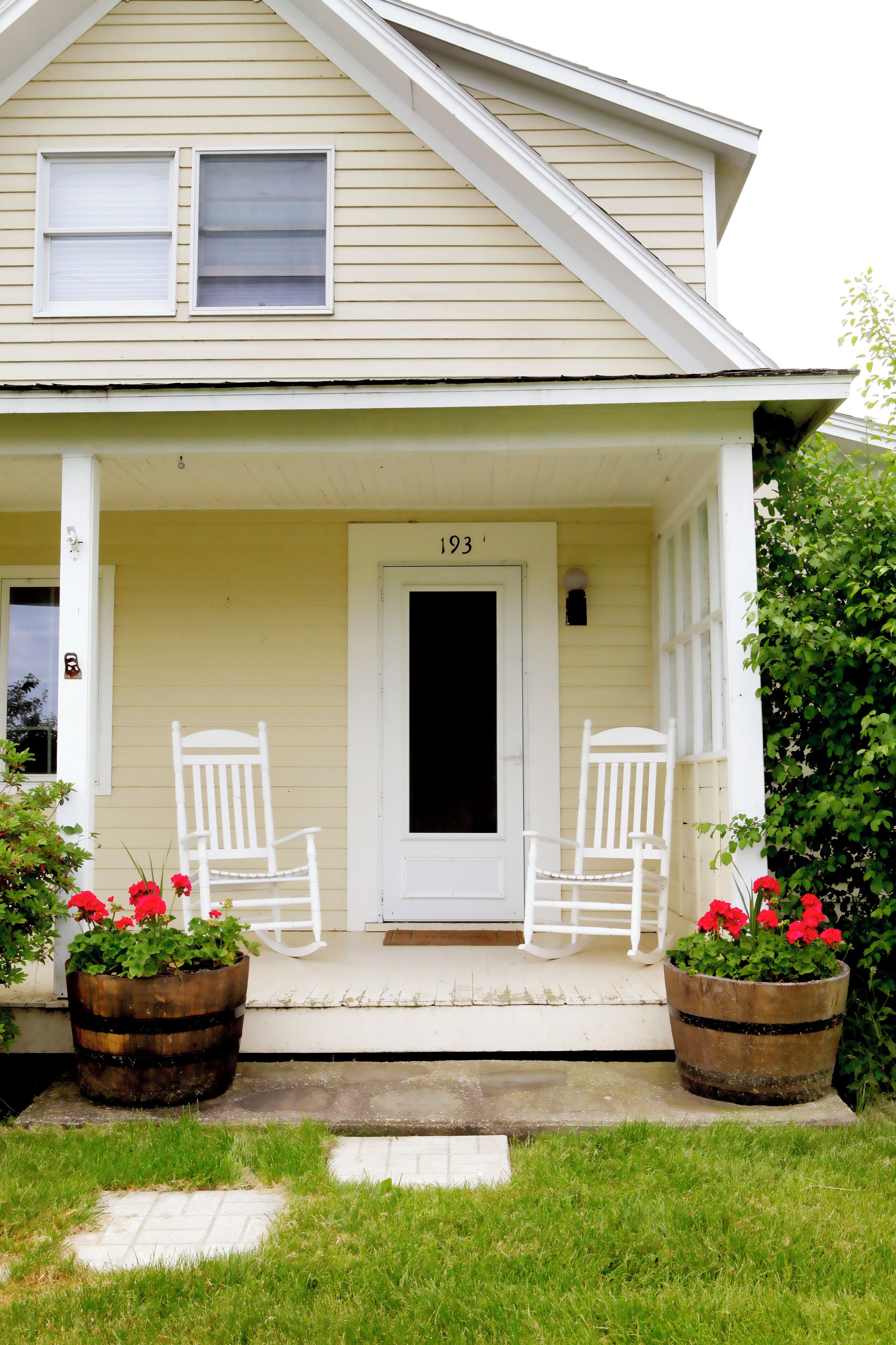 A house with a porch, two white rocking chairs, and two large flower pots with red flowers.