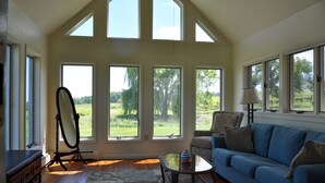 A spacious living room with a blue sofa, a glass coffee table, and a large mirror. There are multiple windows offering views of a green landscape. - The Inn at Glenora Wine Cellars (Dundee)