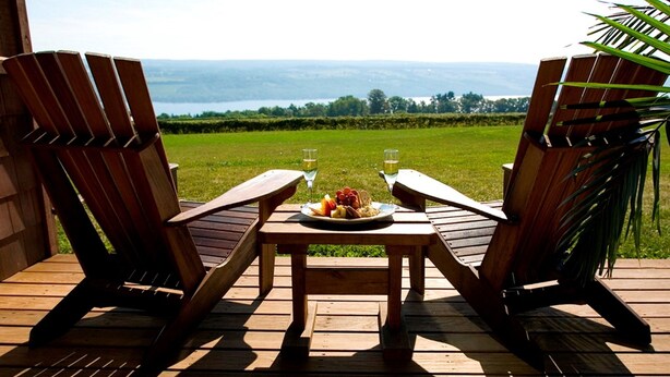 Two wooden chairs facing a table with a plate of fruit and two glasses of water on a wooden deck with a view of a body of water and greenery. - The Inn at Glenora Wine Cellars (Dundee)
