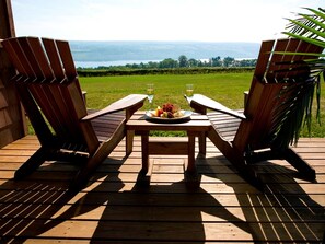 Deux chaises en bois sont placées face à une table sur laquelle se trouve une assiette de fruits et deux verres d’eau. L’ensemble se trouve sur une terrasse en bois, d’où l’on peut admirer un plan d’eau et la verdure environnante.