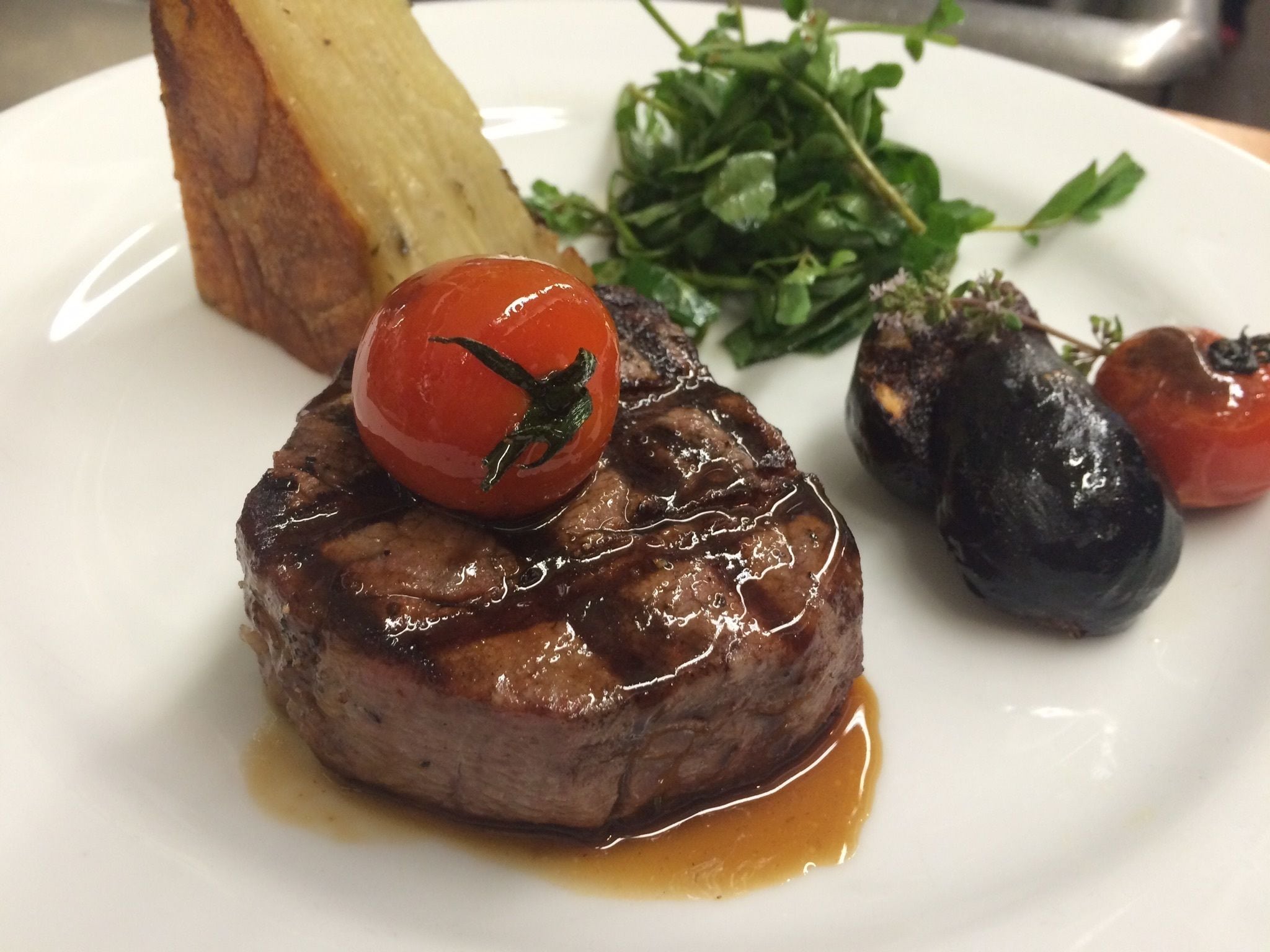 A plate with a steak topped with a cherry tomato, accompanied by roasted vegetables and a piece of bread.