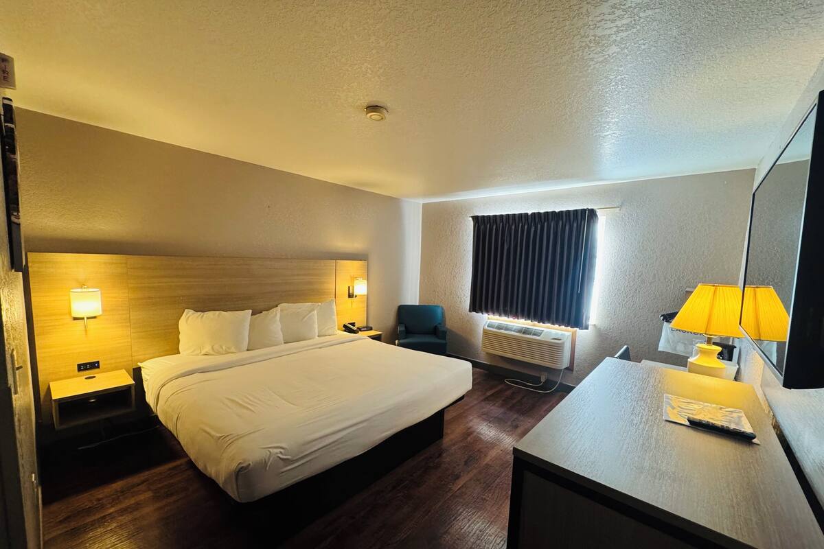 A hotel room with a large bed, bedside tables with lamps, a desk with a lamp, a window with curtains, and a wall-mounted air conditioner.