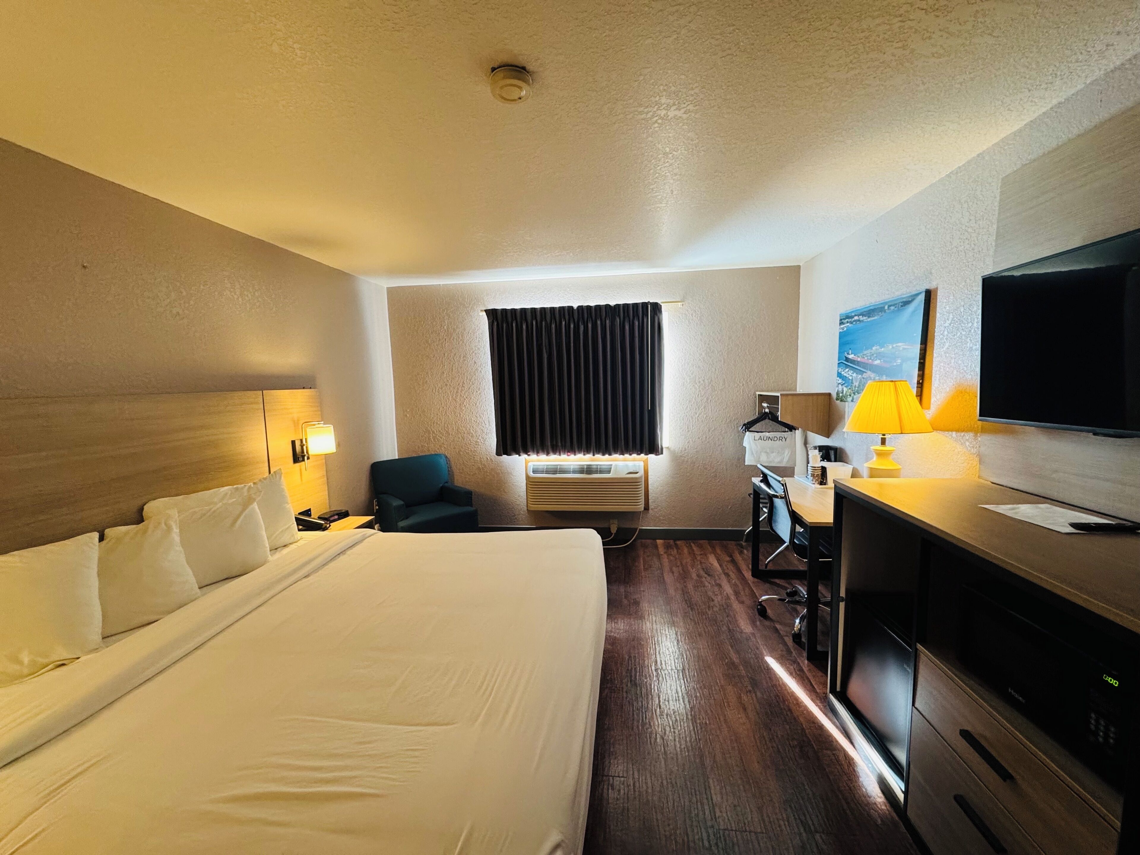 Quarto de hotel com uma cama grande, uma escrivaninha com abajur, uma cadeira, uma TV e uma janela com cortinas.