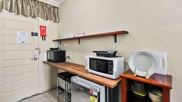 A compact kitchenette with a microwave, toaster, and small refrigerator. There is a shelf above the counter holding dishes and a sign on the door.