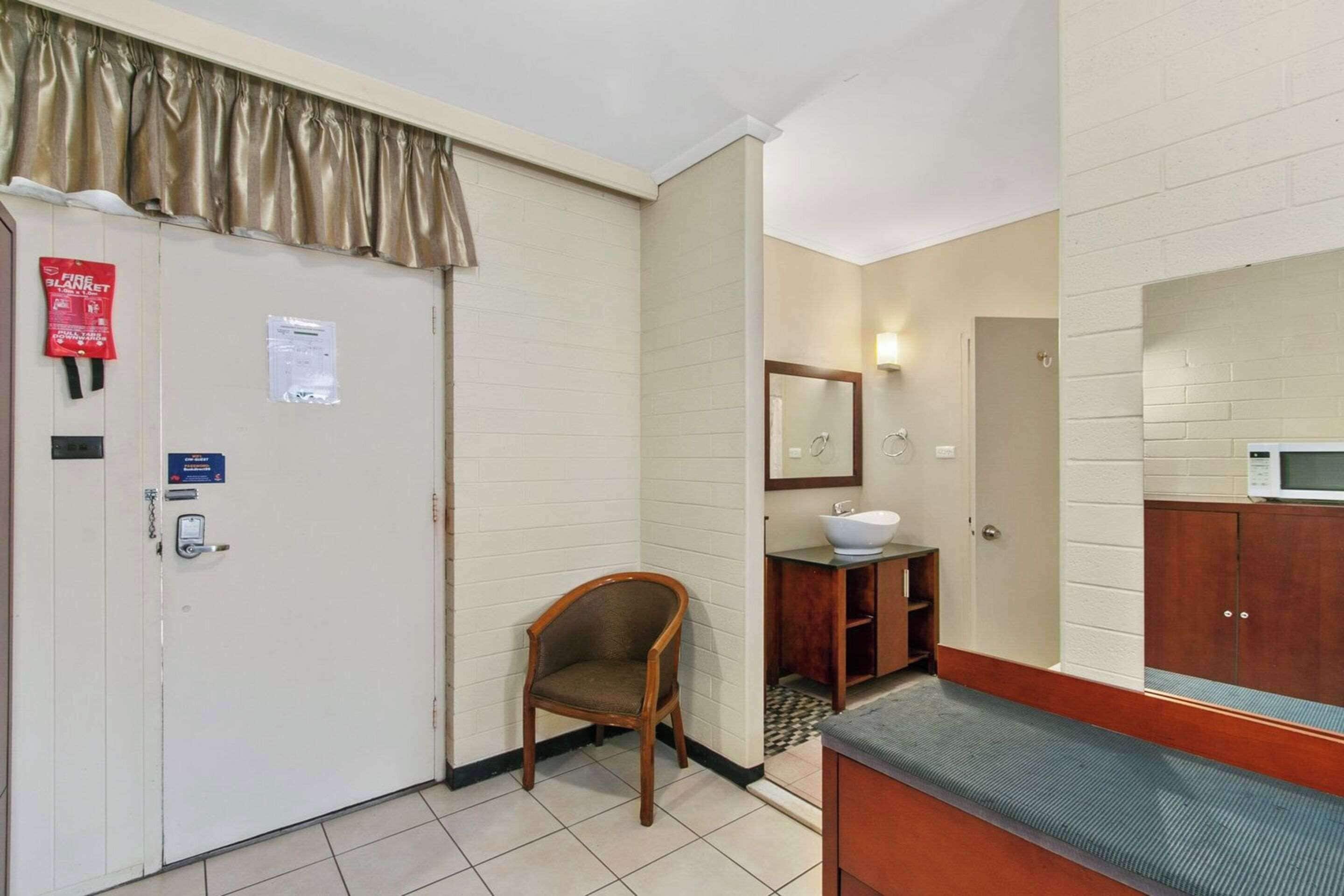 A hotel room with a white door, a wooden chair, a sink, a mirror, and a microwave.