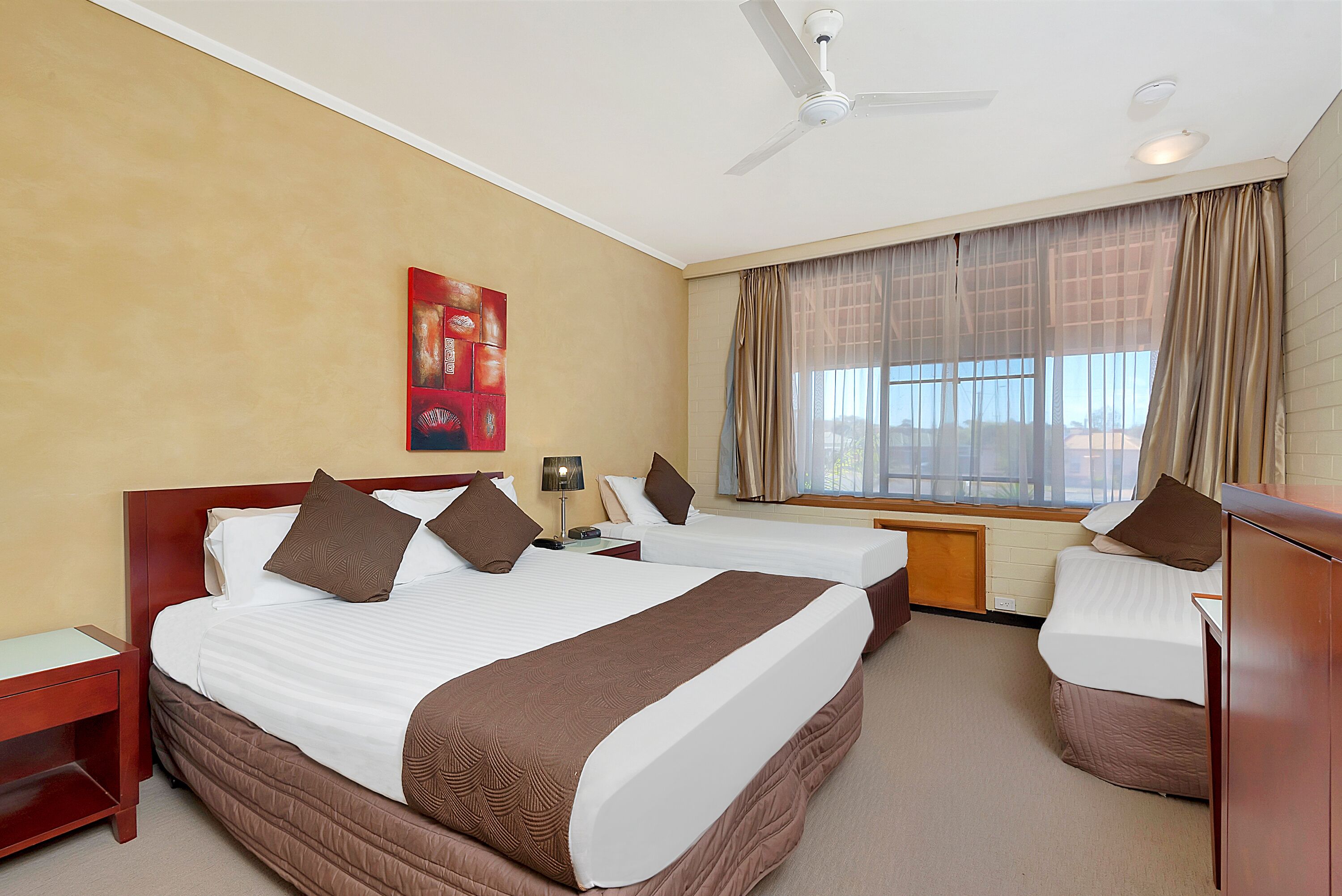 A hotel room with two beds, a ceiling fan, a window with curtains, and a painting on the wall.