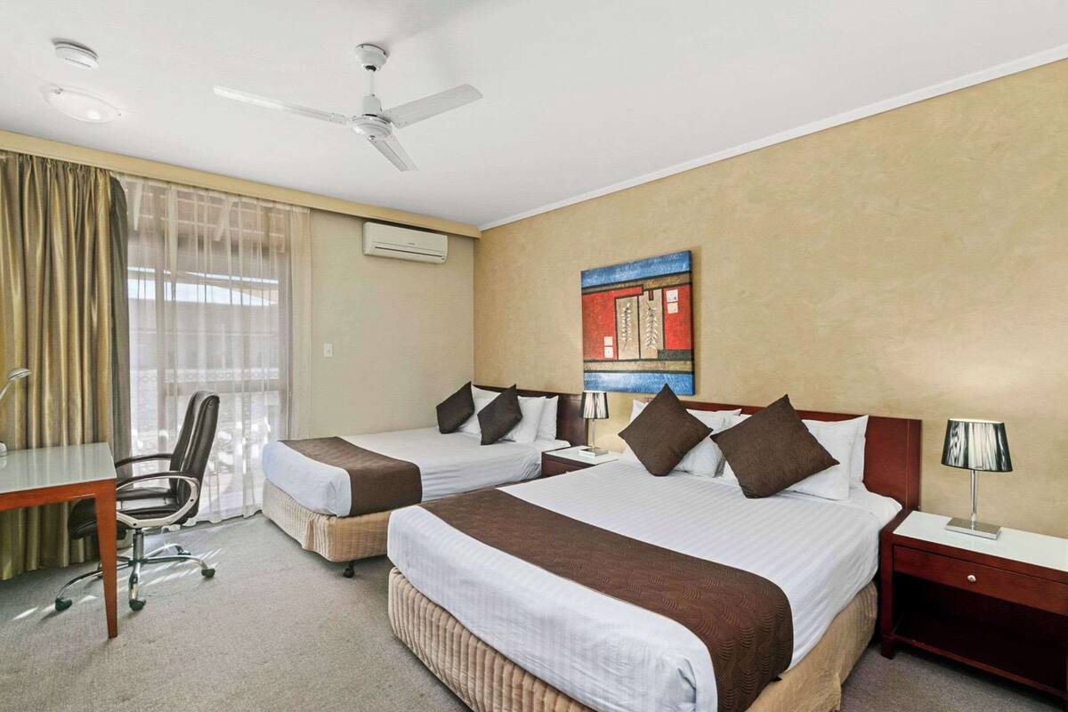 Ein Hotelzimmer mit zwei Betten, einem Schreibtisch, einem Stuhl, einem Deckenventilator und einem Bild an der Wand.
