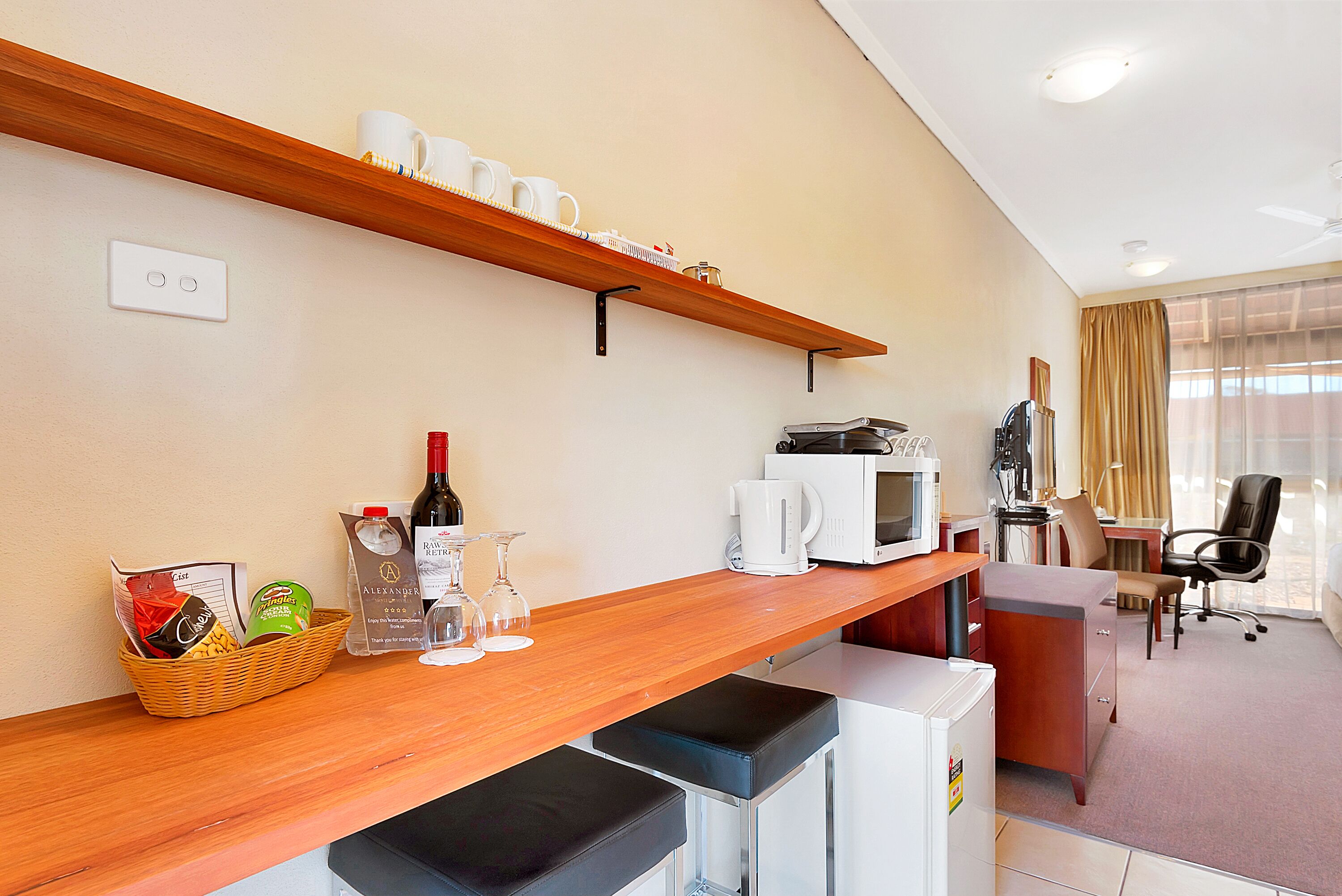 Een hotelkamer met een houten bar, een magnetron, een koffiezetapparaat en een zithoek met een bureau en stoelen.