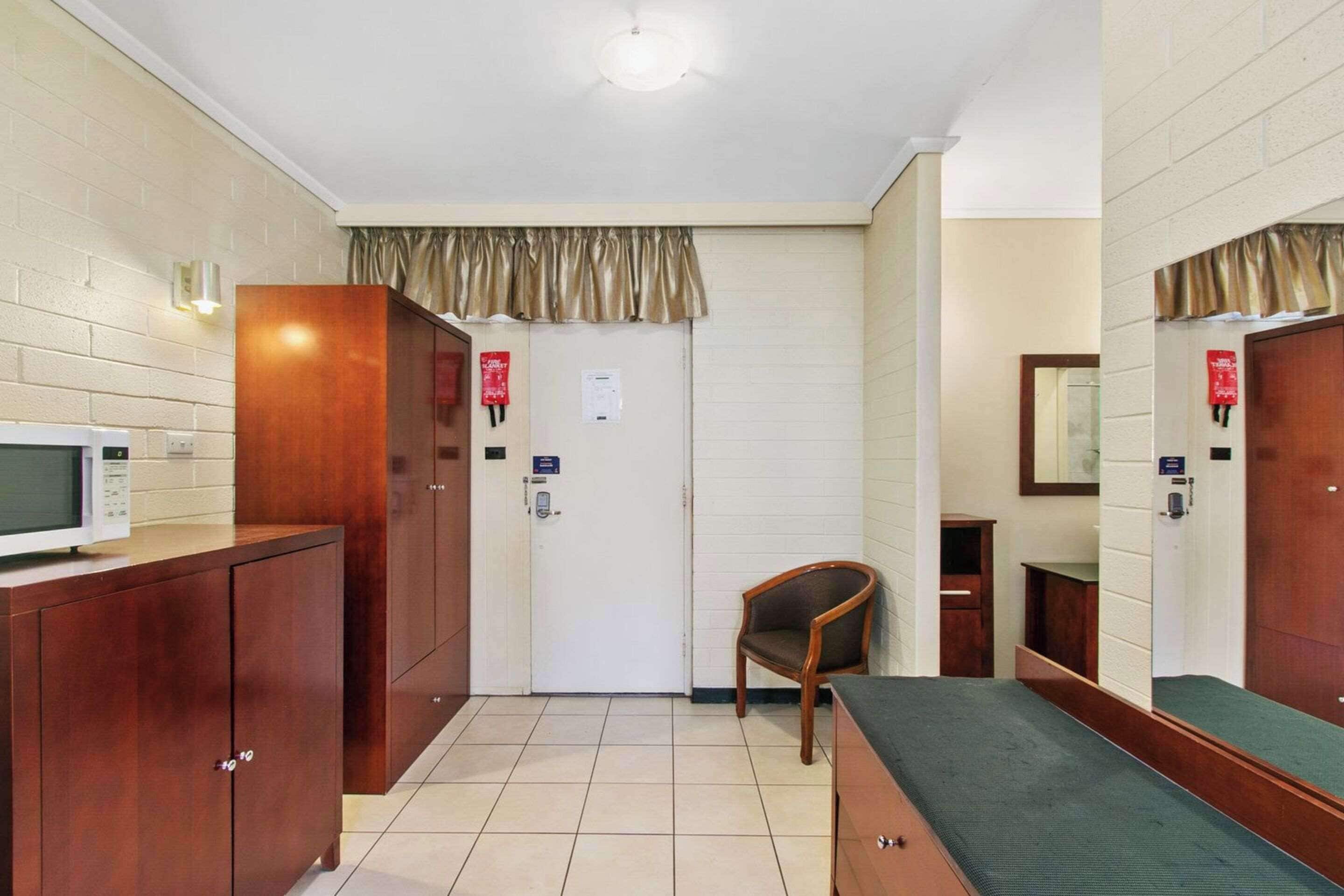 A hotel room with a wooden cabinet, a bed, a chair, a microwave, and a door with a red sign.
