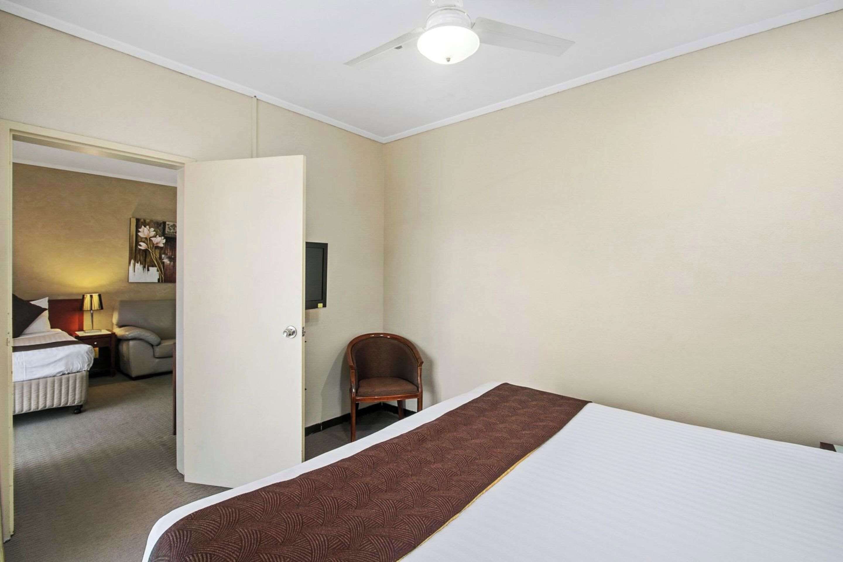 A hotel room with a bed, a chair, a desk with a lamp, and a door leading to another room.