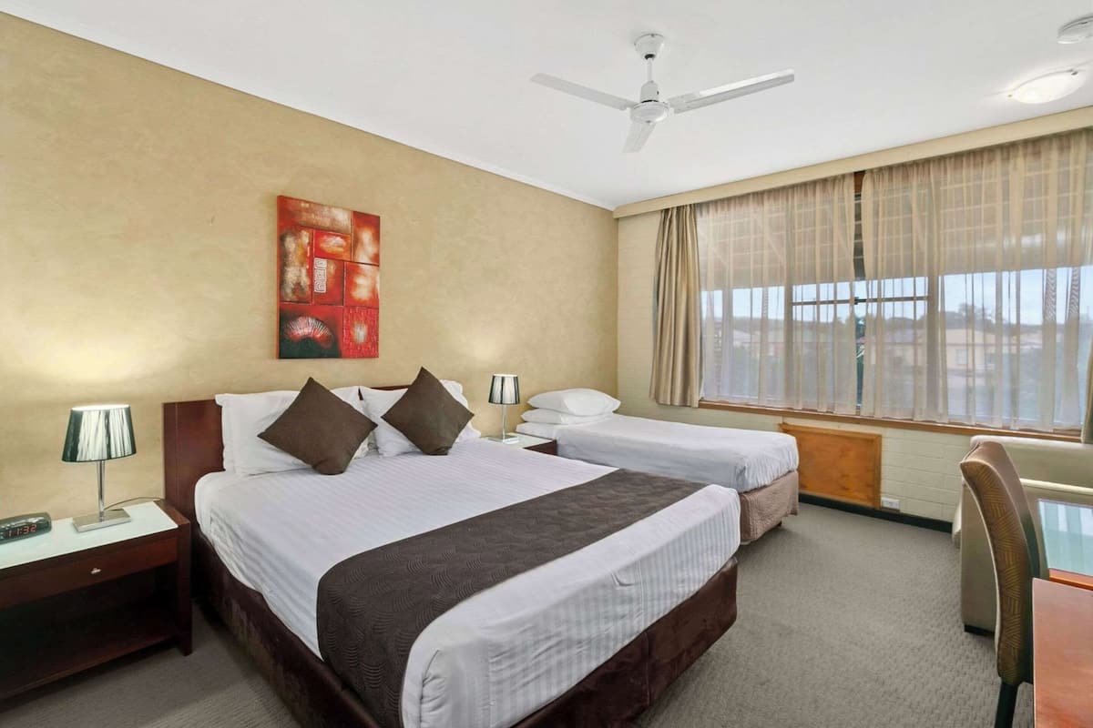 Ein Hotelzimmer mit zwei Betten, einem Schreibtisch, einem Stuhl, einem Deckenventilator und einem großen Fenster mit Vorhängen.