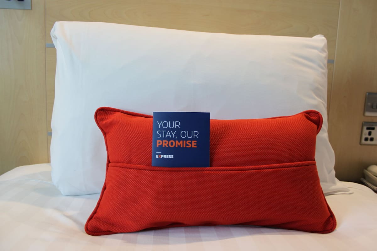 A bed with a white pillow and a red pillow with a tag that reads "YOUR STAY, OUR PROMISE".