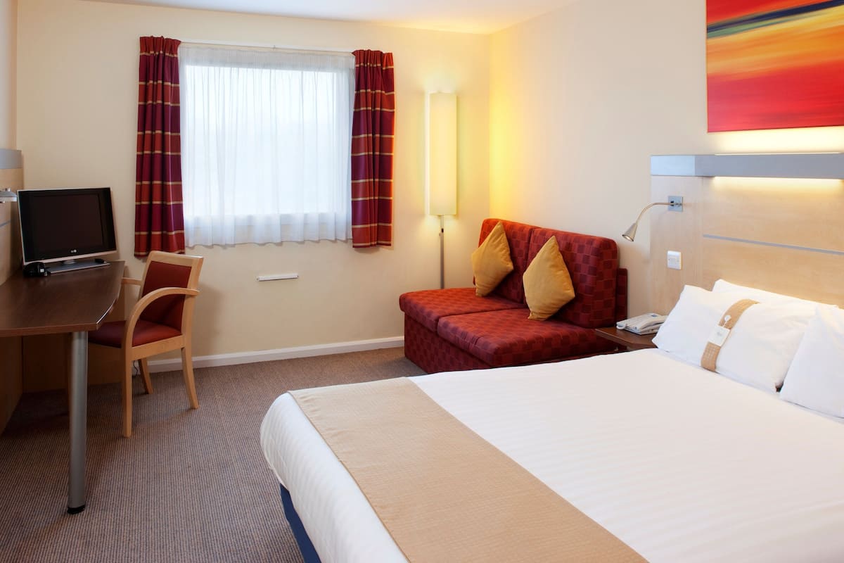 A hotel room with a bed, a desk with a computer, a chair, a sofa, and a lamp.