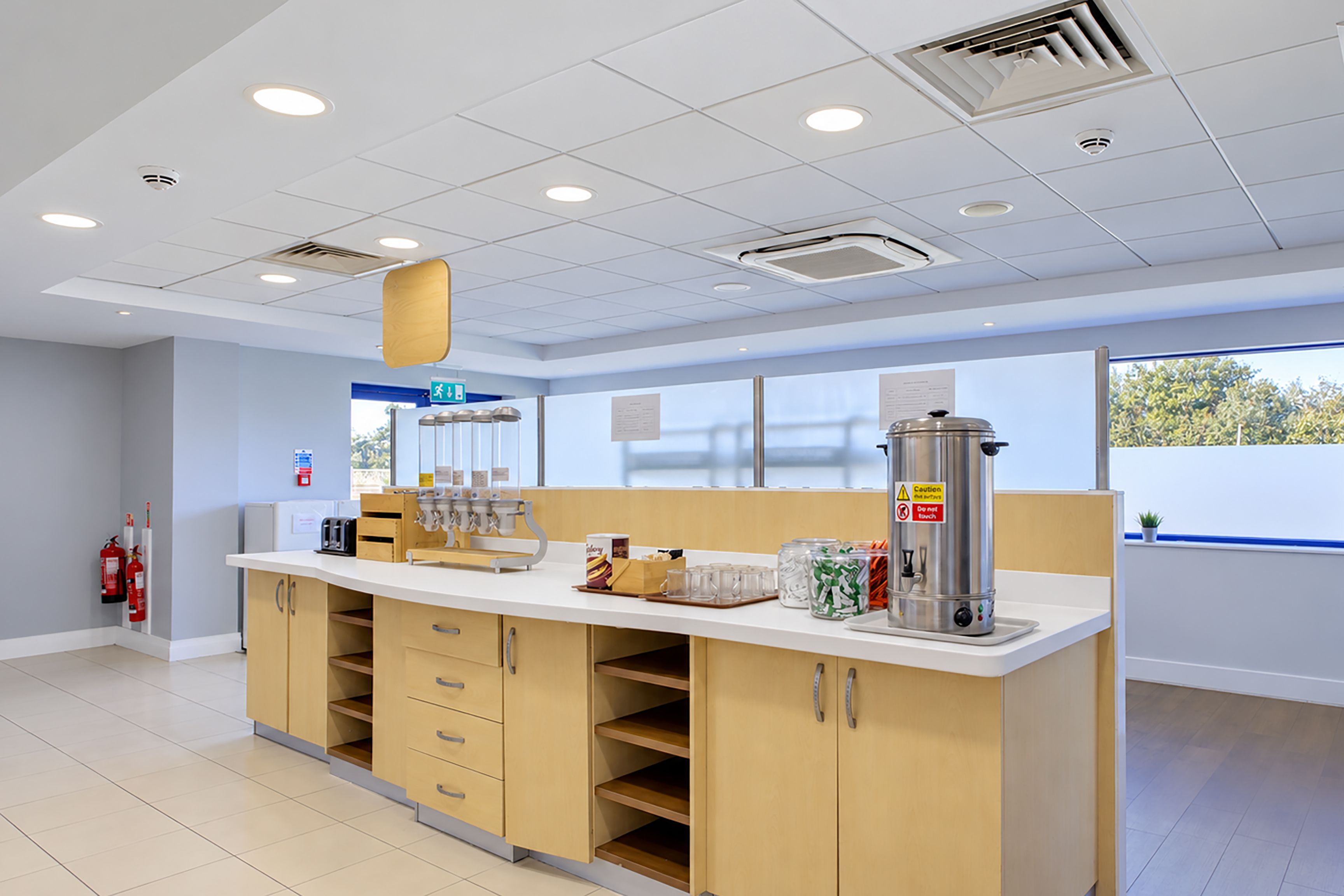 A clean, modern kitchenette with a white countertop, wooden cabinets, and a stainless steel coffee maker.