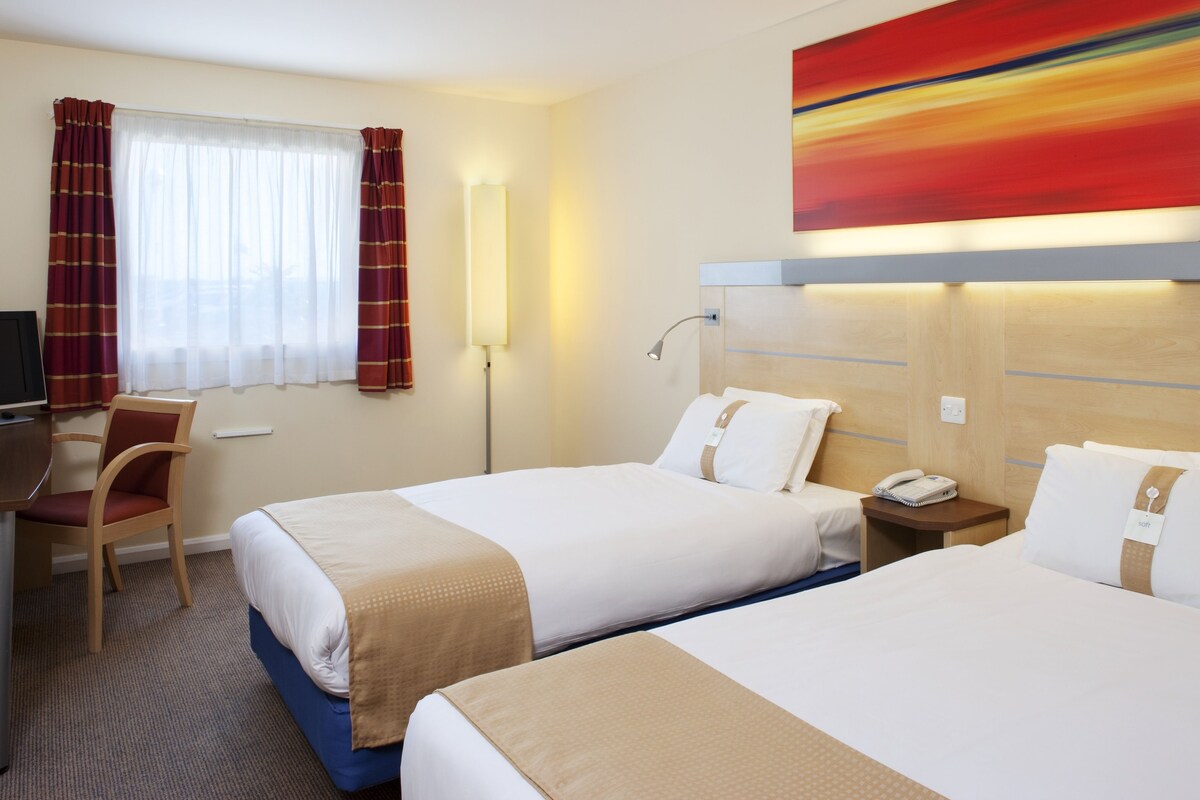 Ett hotellrum med två sängar, ett skrivbord, en stol, en lampa och en tavla på väggen.