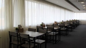 Une salle de conférence avec des rangées de tables et de chaises, de grandes fenêtres ornées de rideaux légers et des luminaires au plafond.