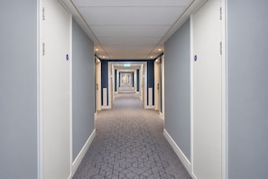 A hotel corridor with a geometric patterned carpet, doors with blue signage, and a ceiling with recessed lighting.