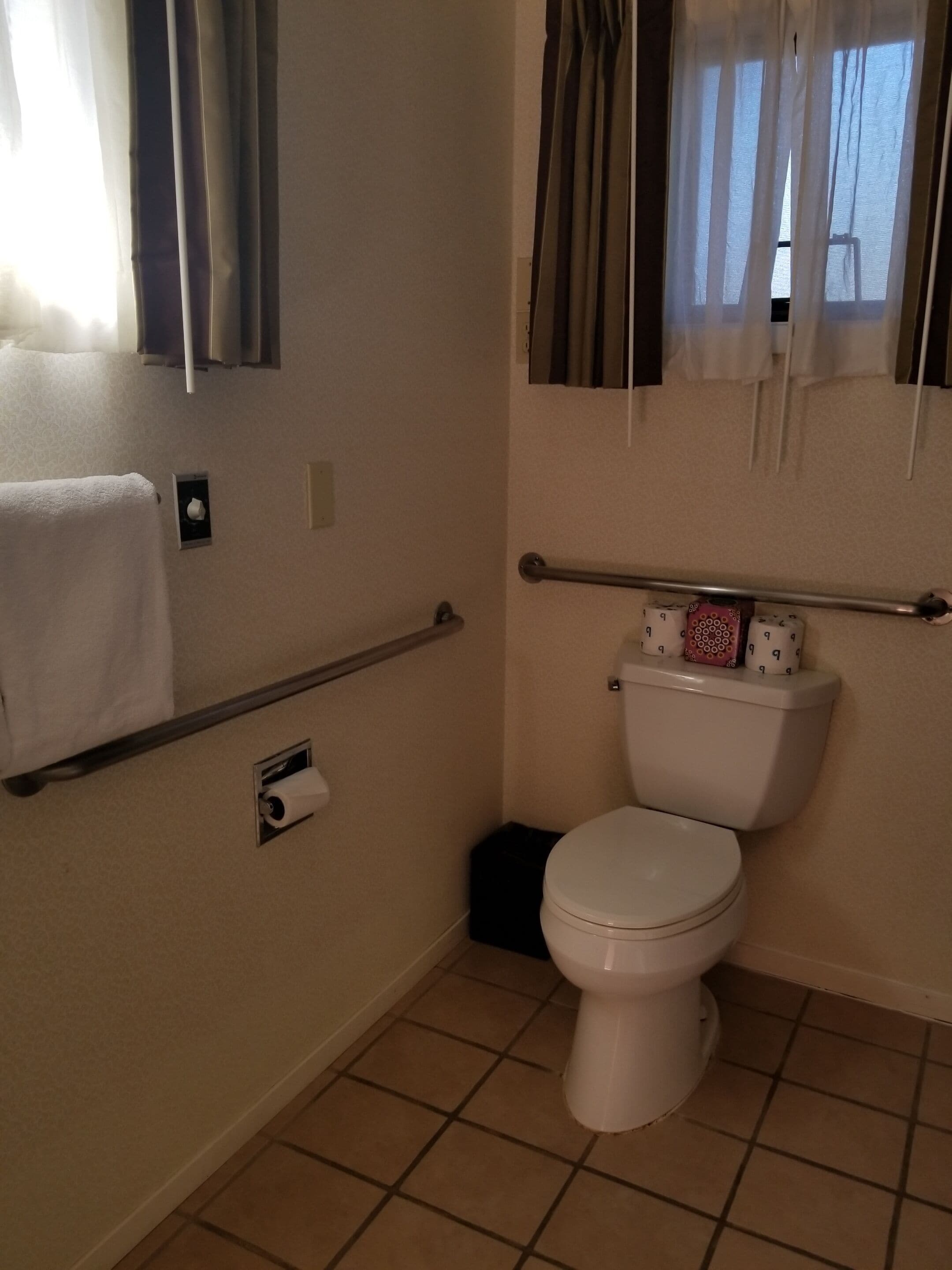 A bathroom with a toilet, a towel rack, a toilet paper holder, and a window with curtains.