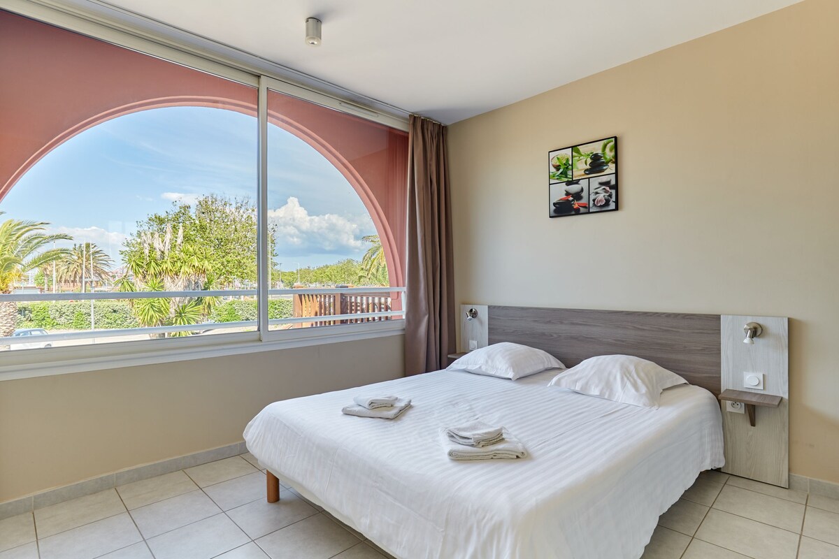 A hotel room with a large window, a bed with white linens, a headboard, and a small wall-mounted nightstand.