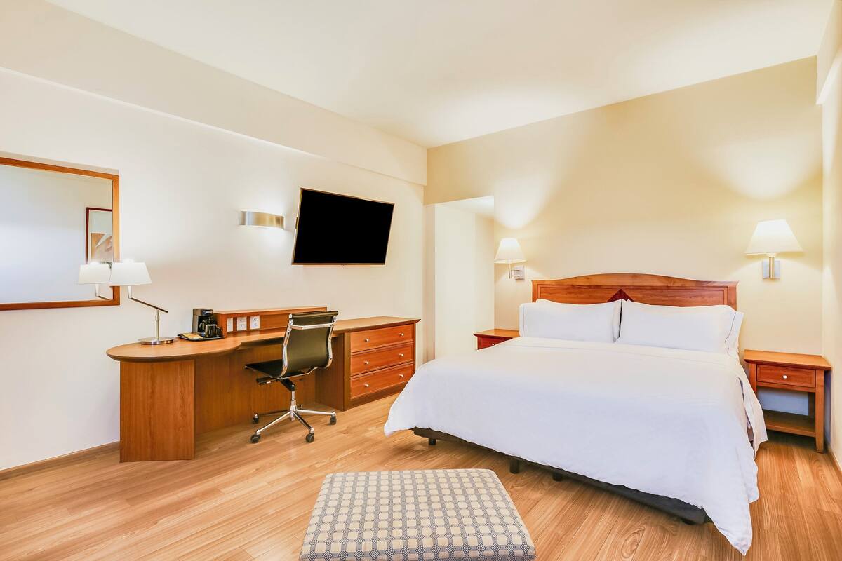 Habitación de hotel con cama, escritorio, silla, televisor y lámparas de noche.