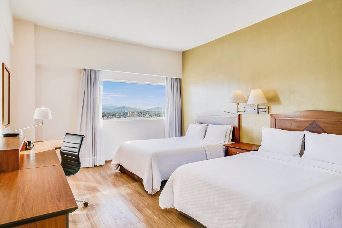 Habitación de hotel con dos camas, un escritorio, una silla y vista a la ciudad.