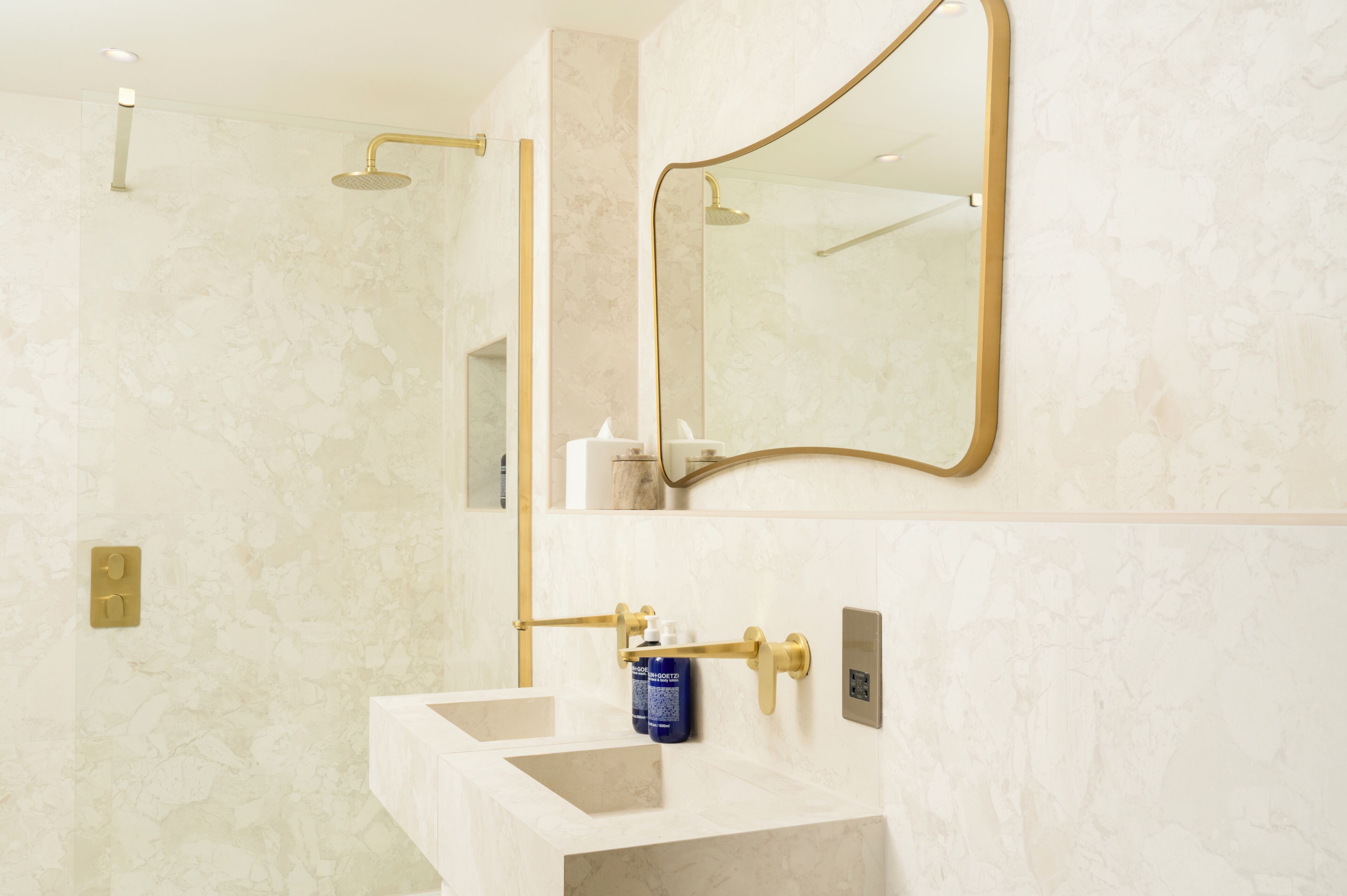 A bathroom with a marble wall, a gold-framed mirror, a white sink, and a gold showerhead.