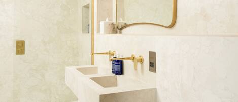 A bathroom with a marble wall, a gold-framed mirror, a white sink, and a gold showerhead.