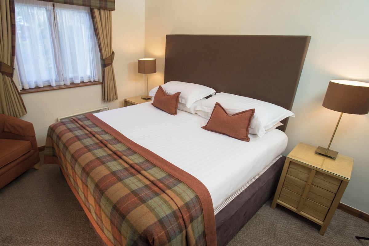 A hotel room with a large bed, a brown armchair, a bedside table with a lamp, and a window with curtains.
