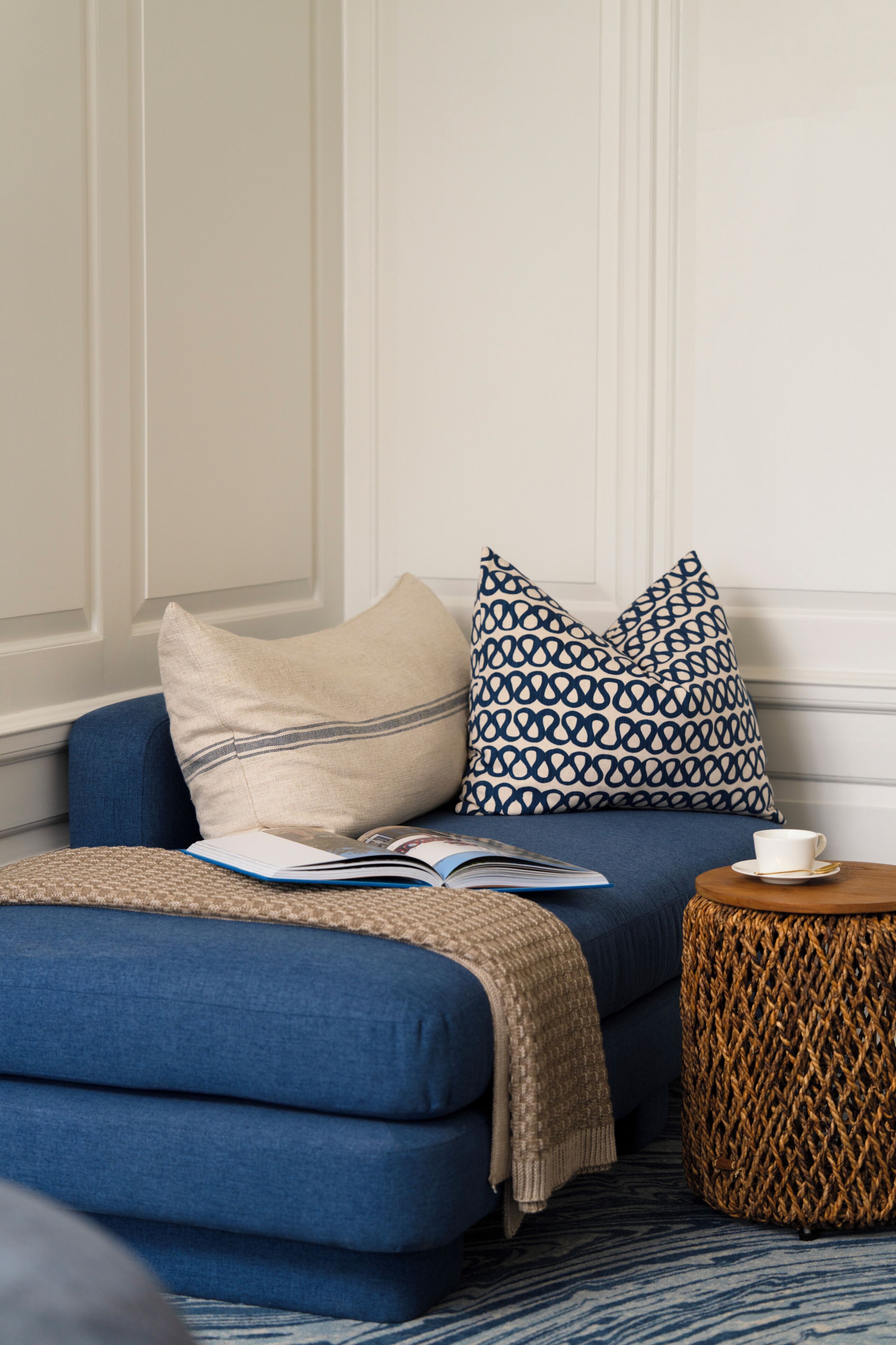 A room with a blue sofa, a patterned pillow, a book, and a small round table with a cup.