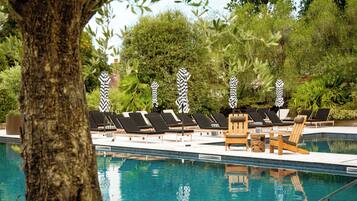 Zona de piscina con sillones de descanso y sombrillas, rodeada de árboles y vegetación.