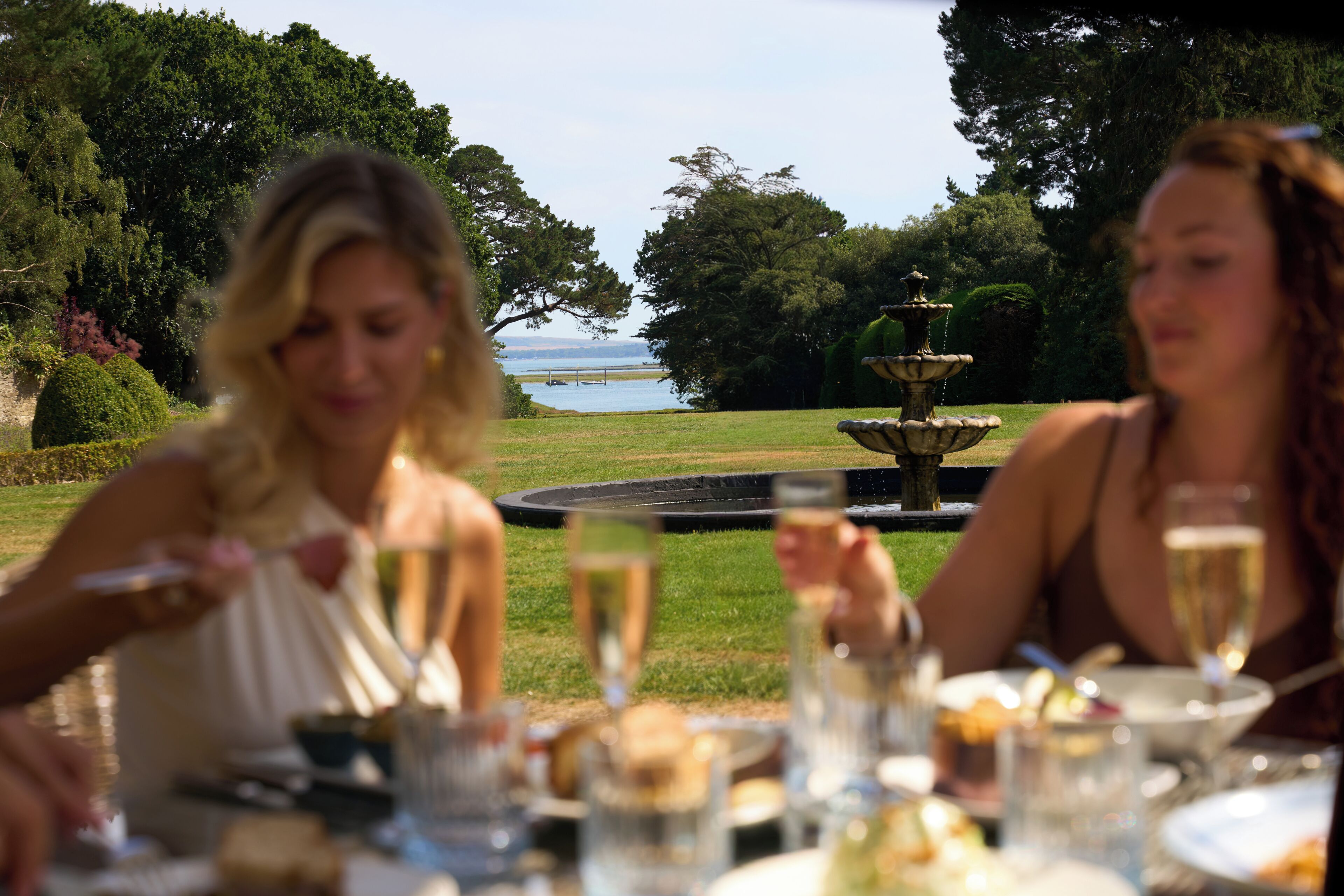 A dining table with food and drinks, a fountain, and a view of the water.