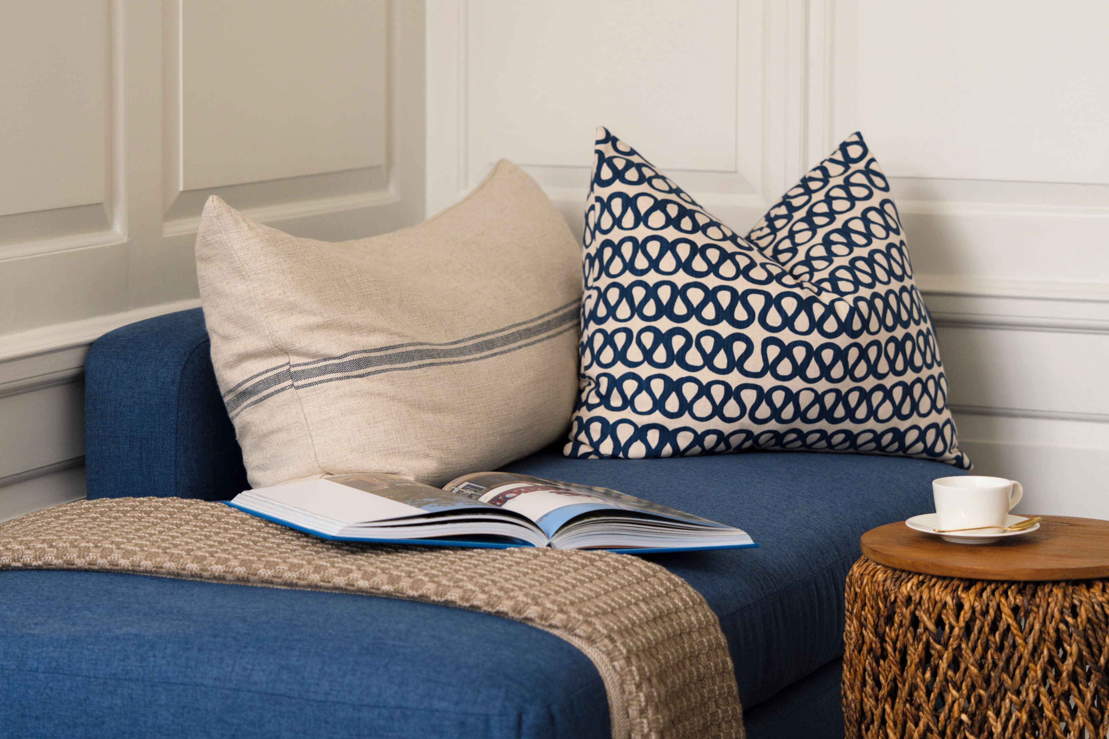 A room with a blue sofa, a patterned pillow, a book, and a small round table with a cup and saucer.