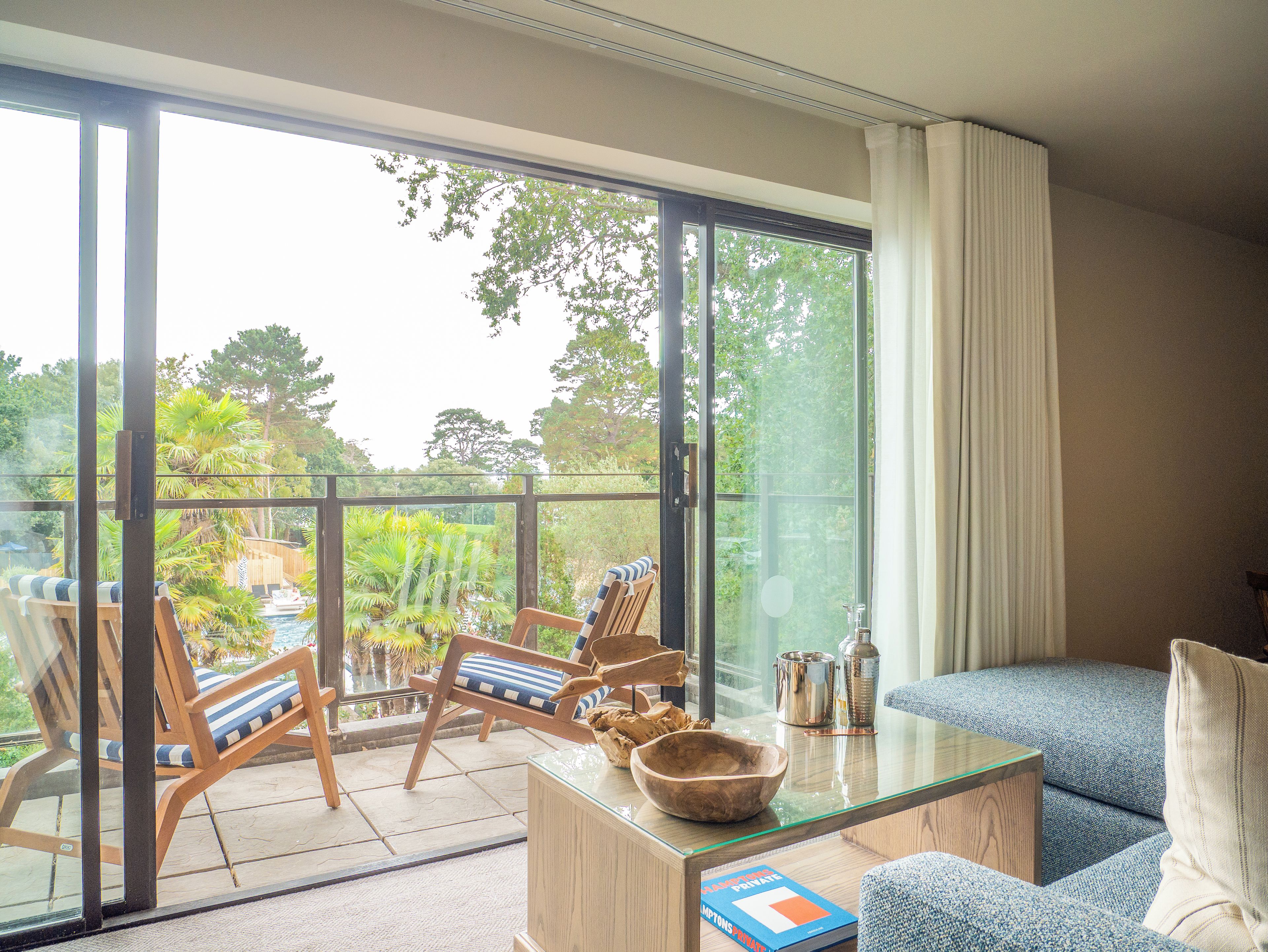A room with a glass table, wooden chairs, a sofa, and a view of greenery outside.