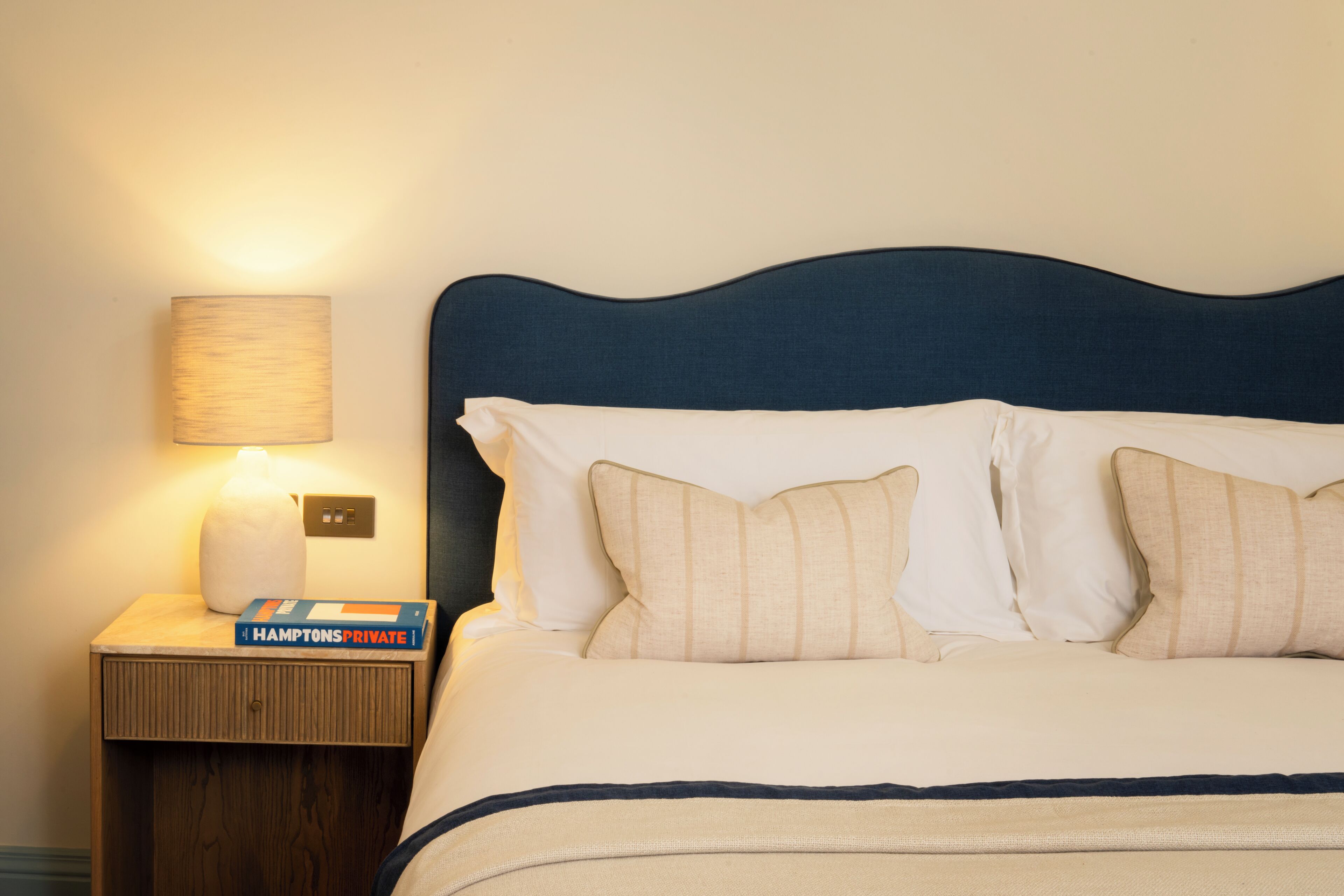 A neatly made bed with a dark headboard, a bedside lamp, and a book titled 'Hampton's Private' on a wooden nightstand.