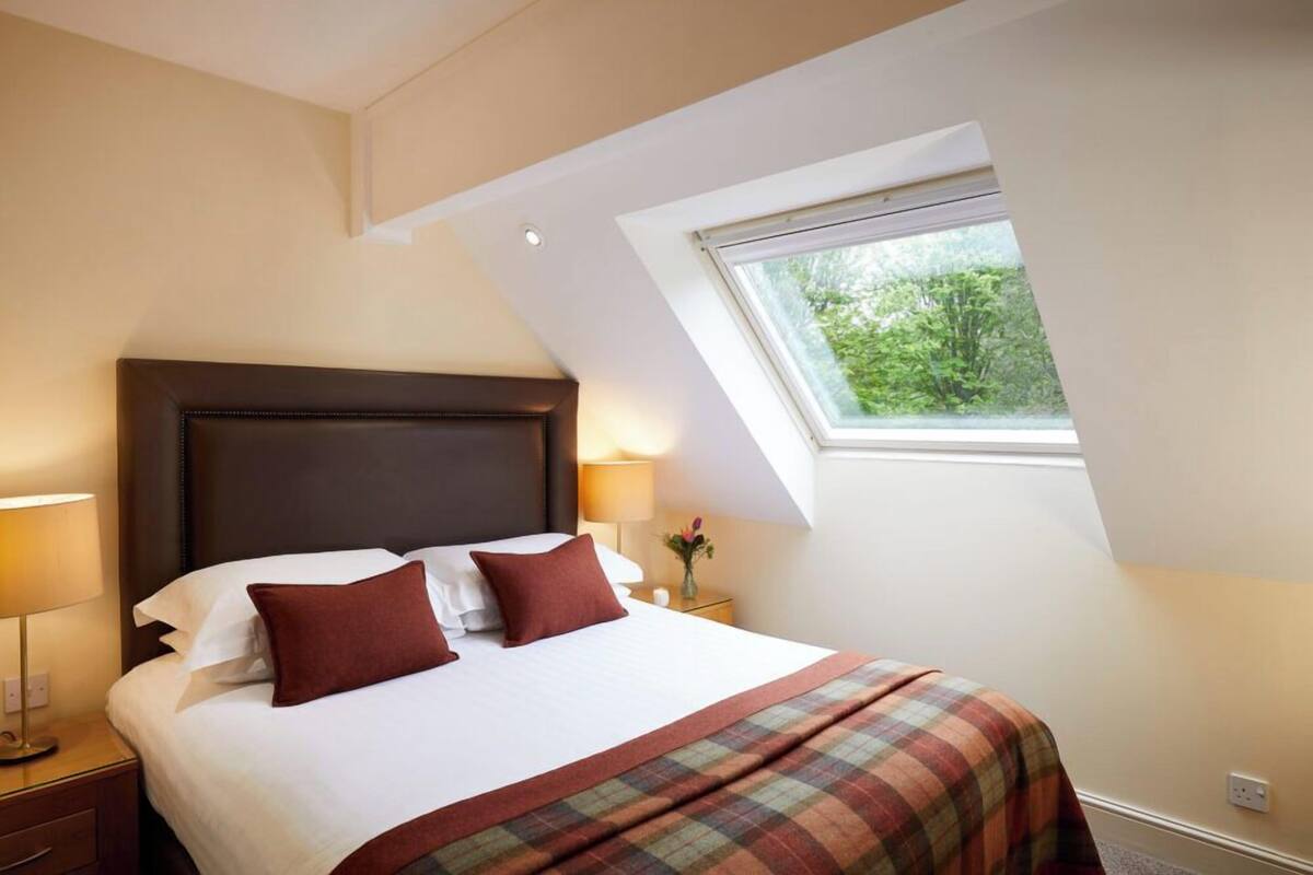 A bedroom with a large bed, two bedside lamps, a window with a view of greenery, and a sloped ceiling.