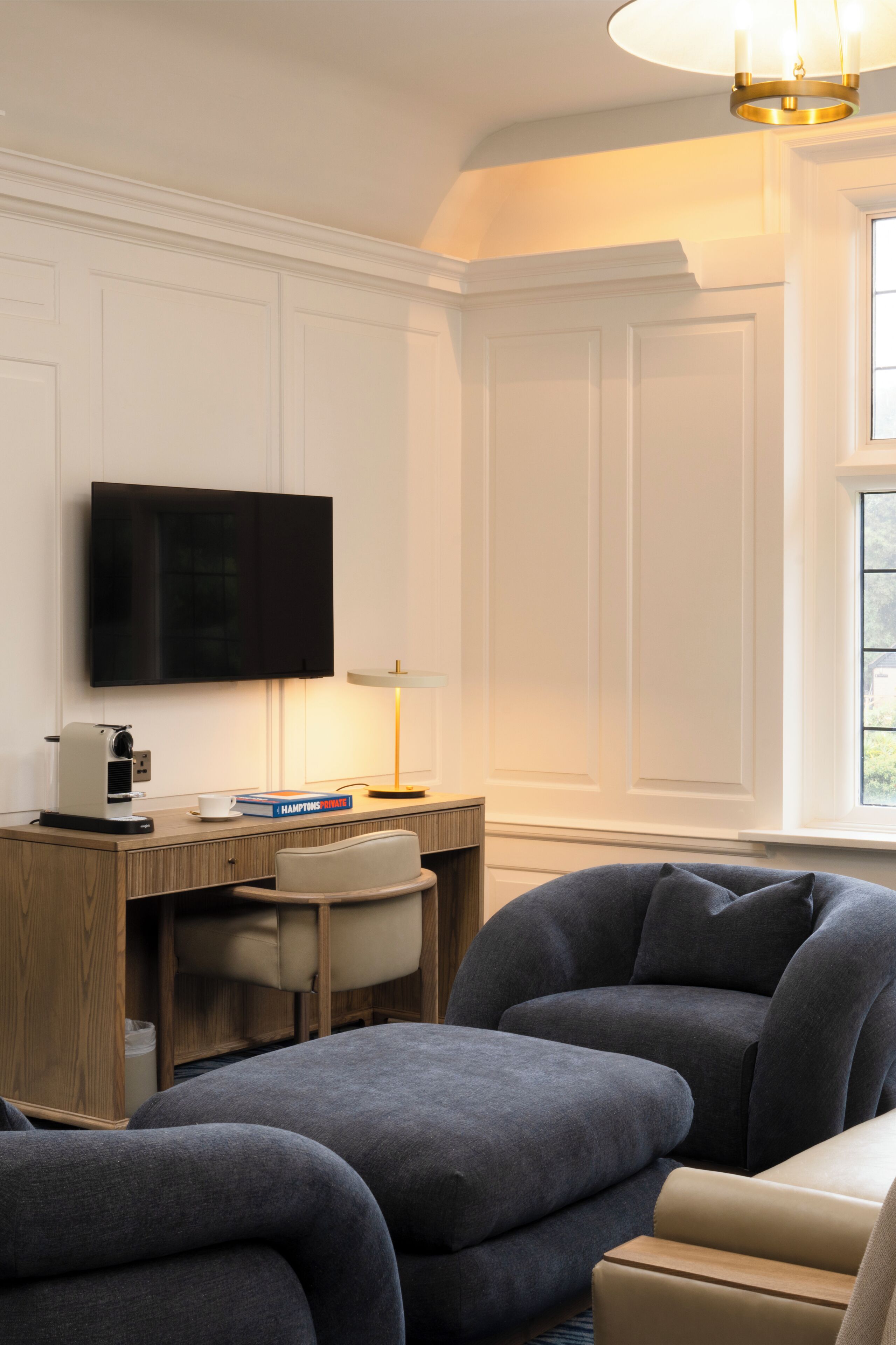 Uma sala de estar moderna com televisão de tela plana na parede, uma escrivaninha de madeira com abajur e uma área de estar confortável com sofás.