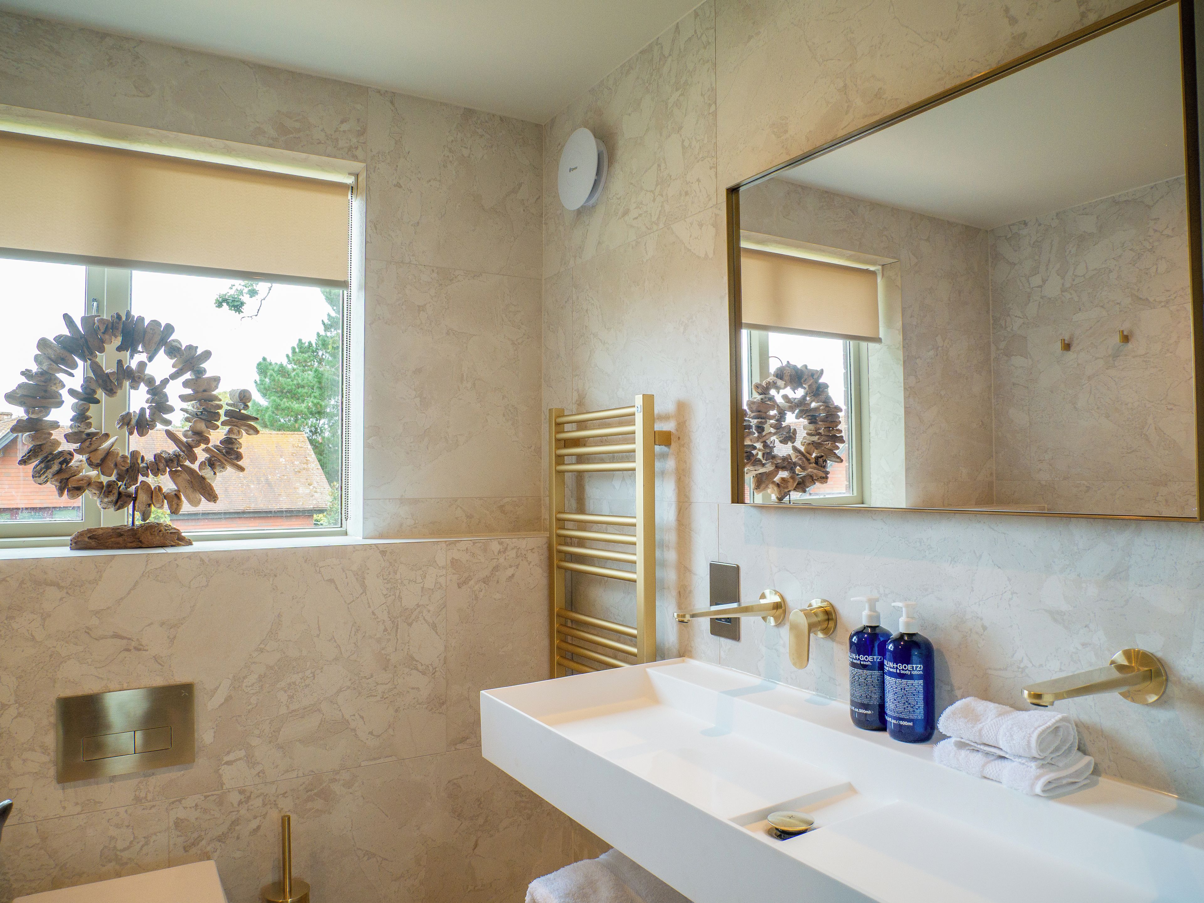 Un baño con lavamanos blanco, detalles dorados, un perchero y una ventana con vista a árboles.