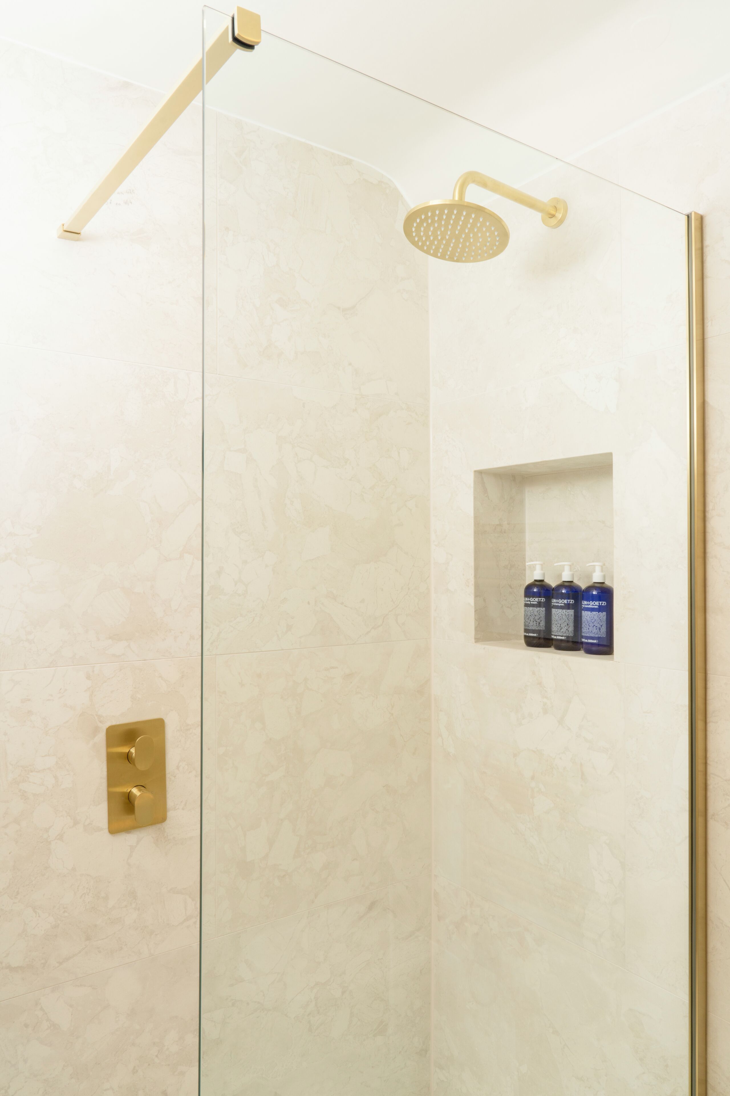 A shower with a glass door, a gold showerhead, and a shelf with toiletries.