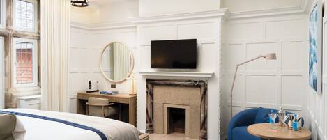 A hotel room with a bed, a desk, a television, a fireplace, a chair, and a small table with drinks.