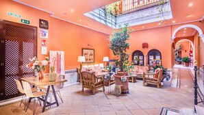 A spacious lobby with a high ceiling, a central seating area with sofas and chairs, a dining table with chairs, and a staircase leading to an upper level. - Hotel Patio de la Alameda (Seville)
