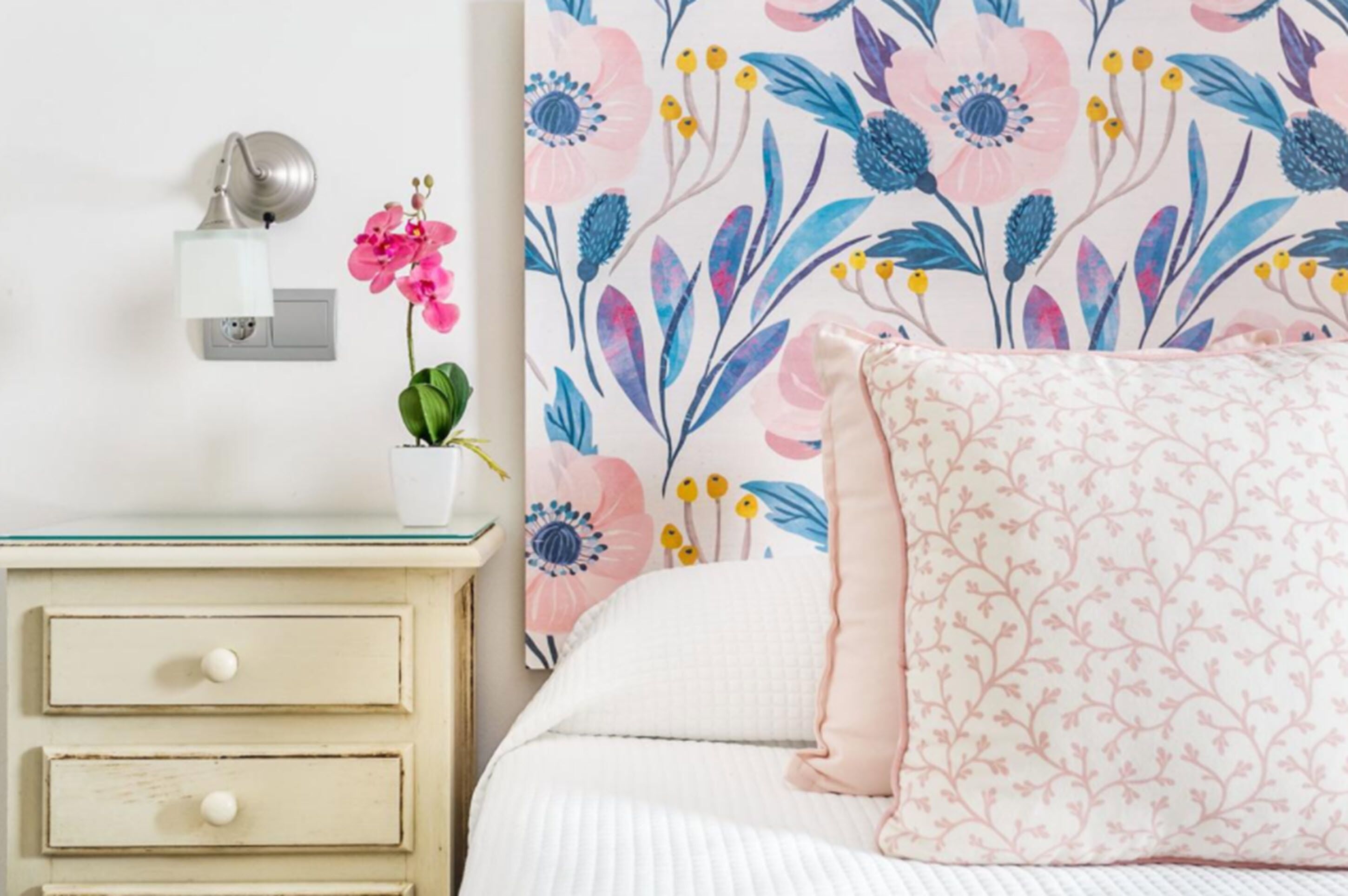A bedroom with a floral wallpaper, a bedside lamp, a nightstand with drawers, and a pink pillow.