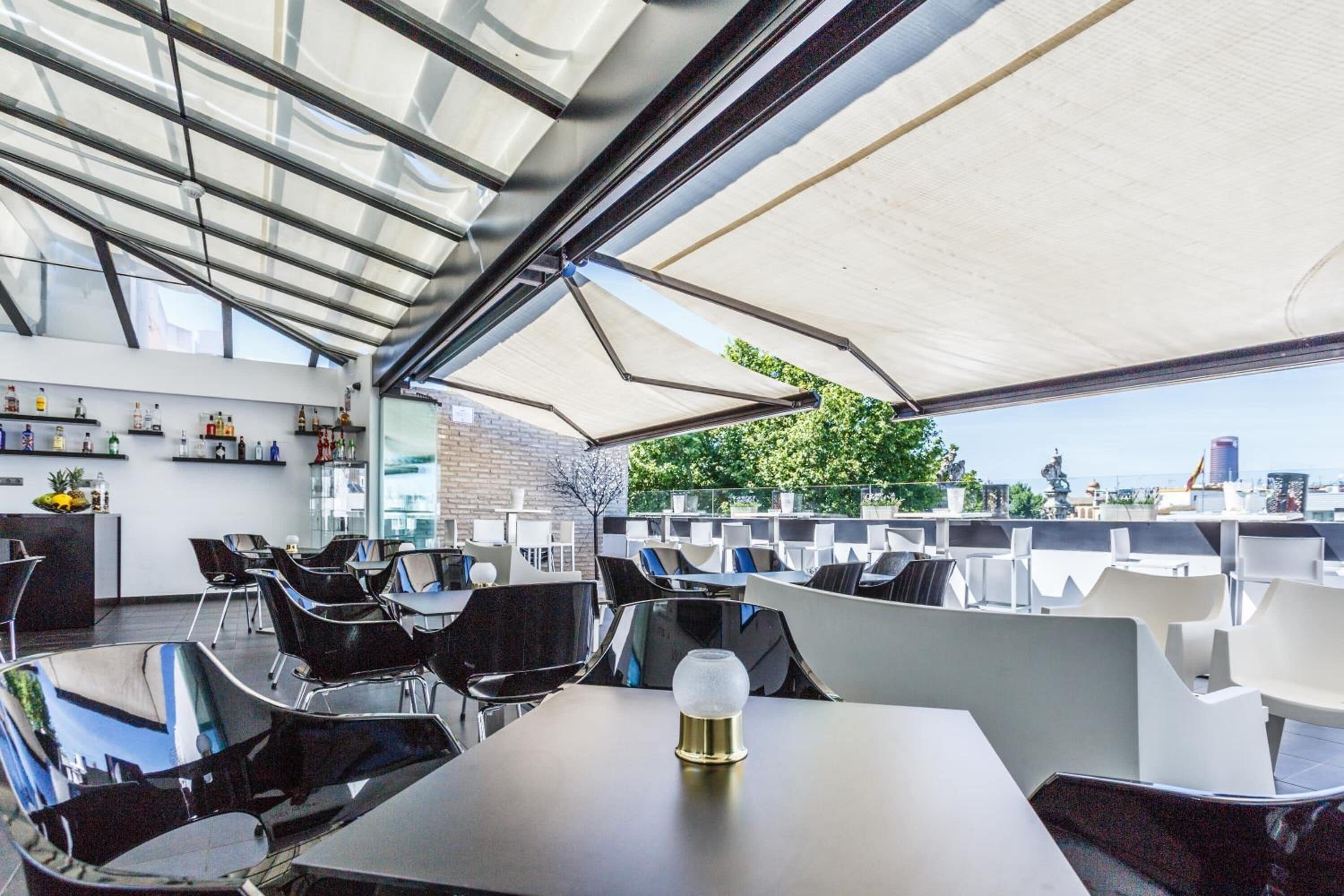 Un espace extérieur aménagé avec des chaises et des tables modernes, un bar où sont exposées différentes bouteilles, et un plafond en verre.