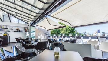Un espace extérieur aménagé avec des chaises et des tables modernes, un bar où sont exposées différentes bouteilles, et un plafond en verre.