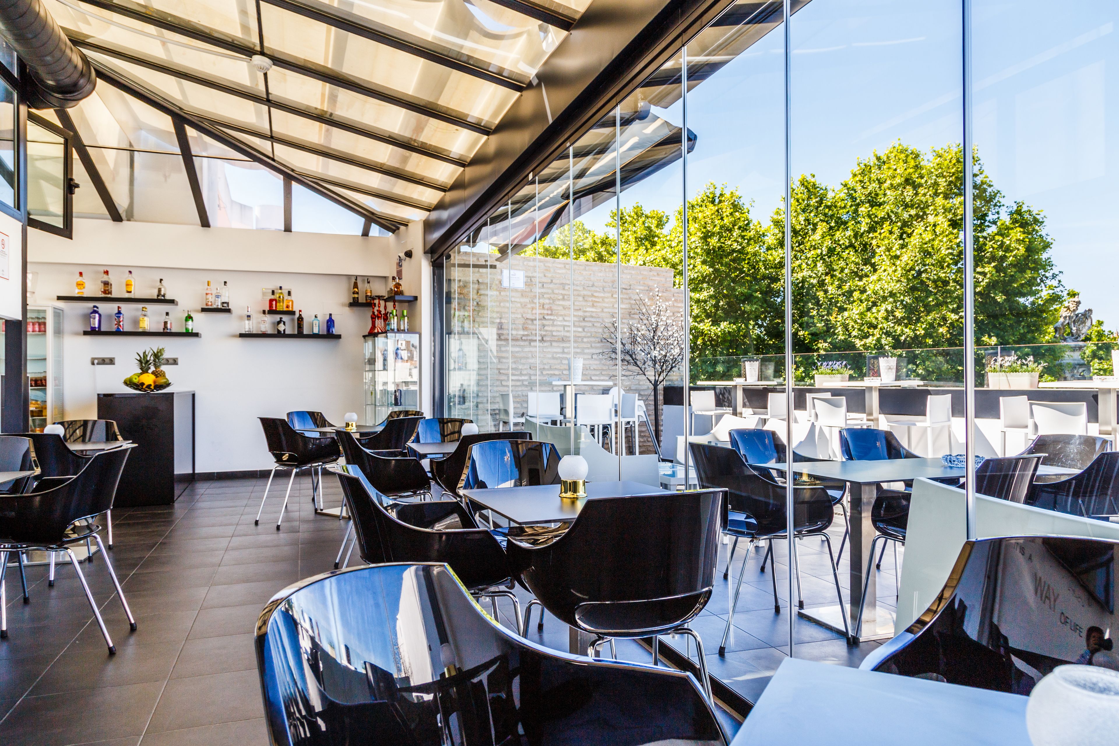 A modern outdoor dining area with glass walls, black chairs, and a variety of beverages on shelves.