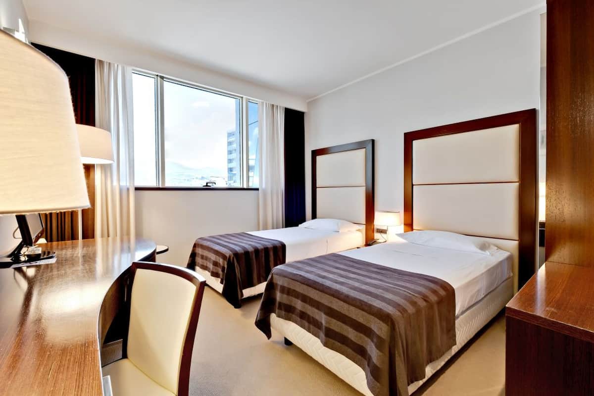 Habitación de hotel con dos camas, un escritorio, una silla, una lámpara y un ventanal con vistas a la ciudad.