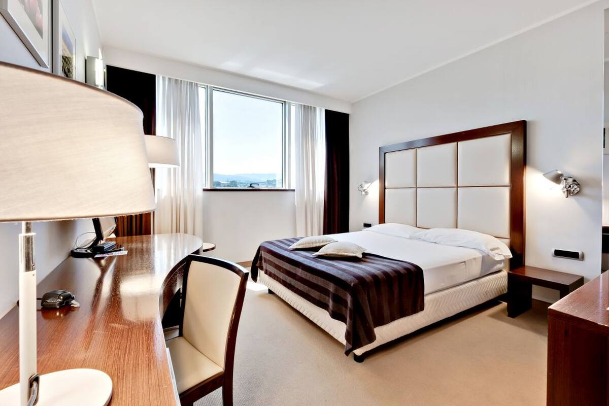 Una habitación de hotel con cama, mesita de noche, lámpara, sillón y escritorio con lámpara.