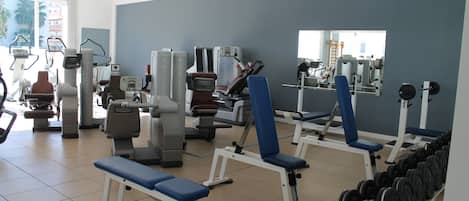 Une salle de sport équipée de divers appareils d’exercice, notamment des bancs et des machines de musculation, et aménagée dans un espace vaste et lumineux grâce à de grandes fenêtres.